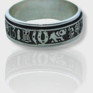Puede incluir: Anillo plateado con una banda negra que presenta varios símbolos de la suerte, como una herradura, un búho y un elefante, además del número 13. El anillo se muestra sobre una superficie blanca, con su reflejo.