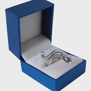 Puede incluir: Un anillo de serpiente plateado con ojos rojos descansa dentro de una caja de joyería azul con interior blanco. El anillo está enrollado en forma de serpiente, con escamas detalladas y una cabeza curvada.