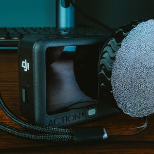 May include: Black action camera with a screen and a textured microphone cover attached. The camera has a wrist strap and the word "ACTION" is visible. The camera is on a wooden surface.