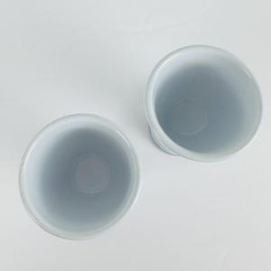 May include: Two cylindrical ceramic cups viewed from directly above. The cups feature a smooth, glossy white interior finish and a light blue exterior glaze. The circular rims are uniform, and the vessels are positioned side by side on a surface.