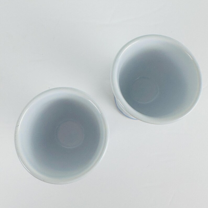 May include: Two cylindrical ceramic cups viewed from directly above. The cups feature a smooth, glossy white interior finish and a light blue exterior glaze. The circular rims are uniform, and the vessels are positioned side by side on a surface.