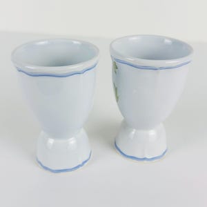 May include: Two light blue ceramic egg cups with a flared rim and a pedestal base. Each cup features a hand-painted wavy blue line detail around the top rim and the base. One cup displays a small green leaf pattern on its side.