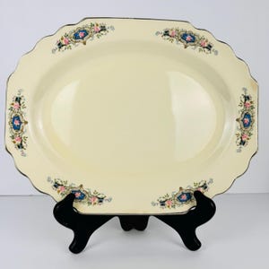 May include: Cream-colored oval serving platter with a scalloped edge and a black stand. The platter features decorative blue and pink floral accents with gold trim. A vintage piece for serving food.