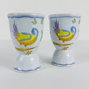May include: Two light blue ceramic egg cups featuring a hand-painted design of a yellow and blue bird with purple tail feathers perched on a brown branch. The cups have a scalloped rim and base, accented with a thin blue painted line detail.