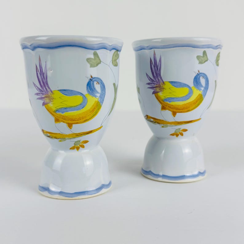 May include: Two light blue ceramic egg cups featuring a hand-painted design of a yellow and blue bird with purple tail feathers perched on a brown branch. The cups have a scalloped rim and base, accented with a thin blue painted line detail.