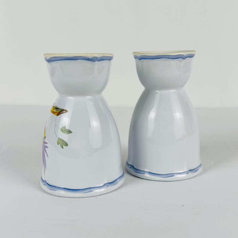 May include: Two light blue ceramic egg cups with a flared rim and base. Each cup features a hand-painted blue trim along the edges. One cup displays a small, colorful floral illustration with green leaves and purple and yellow petals on its side.
