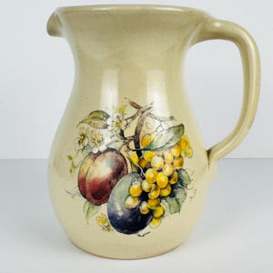 May include: Cream-colored ceramic pitcher with a curved handle and spout. The pitcher is decorated with a colorful illustration of fruit, including grapes, plums, and a peach. The pitcher is likely used for serving beverages.
