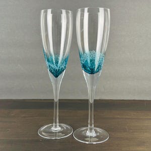 May include: Two clear champagne flutes with a teal, textured design at the base. The flutes have long, slender stems and round bases. The background is a neutral gray, and the flutes are set on a dark wooden surface.