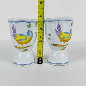 May include: Two white ceramic egg cups featuring hand-painted yellow and blue birds perched on branches with purple tail feathers. A yellow measuring tape is positioned between the cups, showing a height of approximately 3.5 inches.