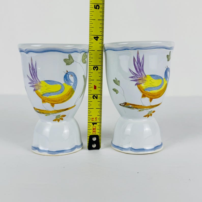 May include: Two white ceramic egg cups featuring hand-painted yellow and blue birds perched on branches with purple tail feathers. A yellow measuring tape is positioned between the cups, showing a height of approximately 3.5 inches.