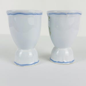 May include: Two light blue ceramic egg cups with a flared top and a pedestal base. Each cup features a hand-painted wavy blue line along the rim and the base. The right egg cup displays a small green leaf pattern on its side, while the left is plain.