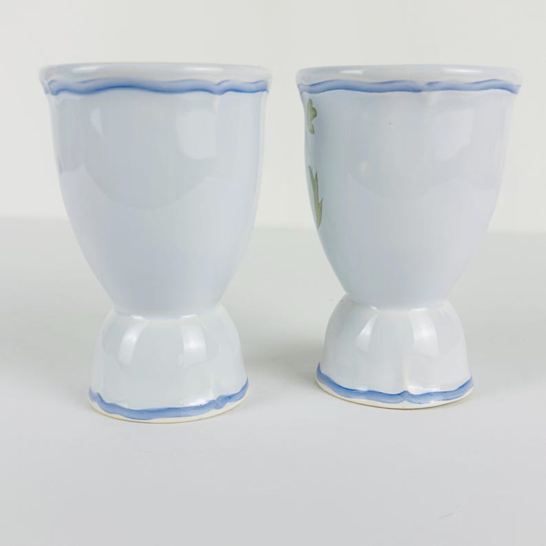 May include: Two light blue ceramic egg cups with a flared top and a pedestal base. Each cup features a hand-painted wavy blue line along the rim and the base. The right egg cup displays a small green leaf pattern on its side, while the left is plain.