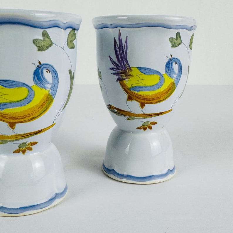 May include: Two ceramic egg cups featuring a hand-painted design of a yellow and blue bird perched on a brown branch. The cups have a light blue base color with a scalloped blue rim and green leaf accents painted around the bird figures.