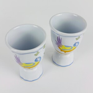 May include: Two white ceramic egg cups featuring a hand-painted design of a bird with a yellow body, blue head, and purple tail feathers. Each cup has a flared rim and a pedestal base with light blue painted trim around the edges and base.