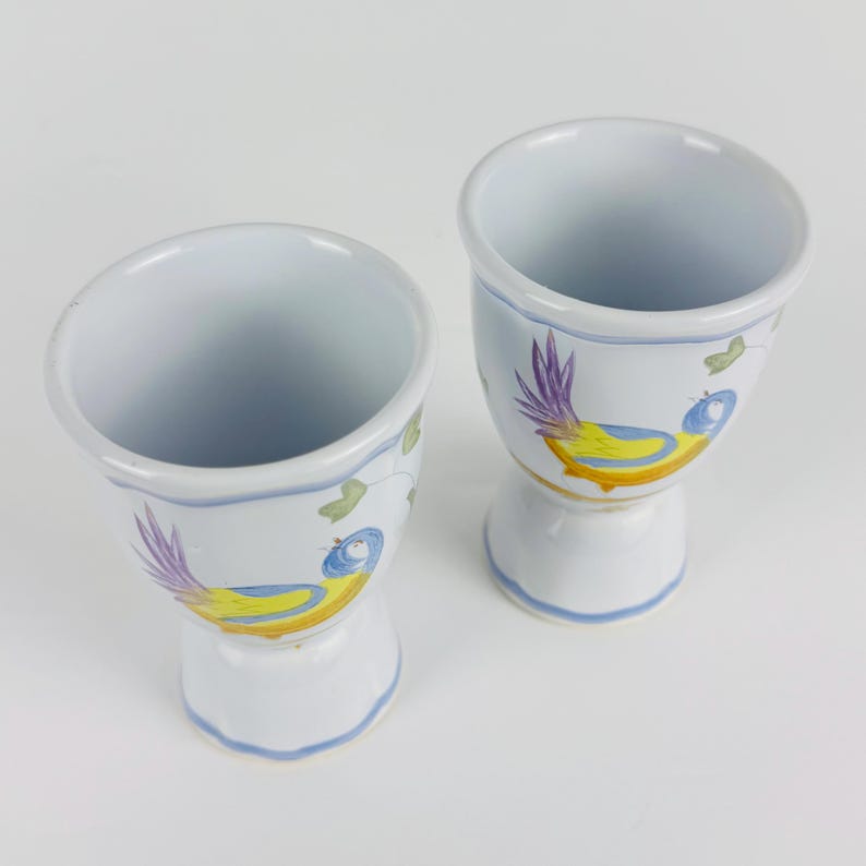 May include: Two white ceramic egg cups featuring a hand-painted design of a bird with a yellow body, blue head, and purple tail feathers. Each cup has a flared rim and a pedestal base with light blue painted trim around the edges and base.
