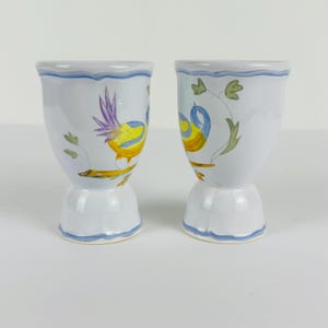 May include: Two white ceramic egg cups featuring hand-painted illustrations of birds perched on branches. The birds are colored in yellow and blue with purple accents. Each cup has a scalloped rim and base, both decorated with a thin blue painted line.