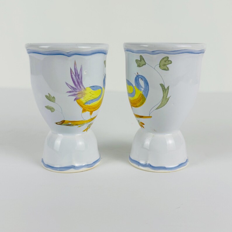 May include: Two white ceramic egg cups featuring hand-painted illustrations of birds perched on branches. The birds are colored in yellow and blue with purple accents. Each cup has a scalloped rim and base, both decorated with a thin blue painted line.
