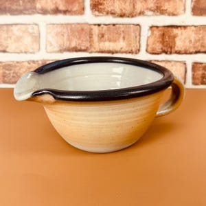 May include: A ceramic creamer pitcher with a light tan exterior and a smooth, off-white interior. The vessel features a dark brown rim that extends to the pouring spout and a small handle on the side, showcasing a simple, functional design.