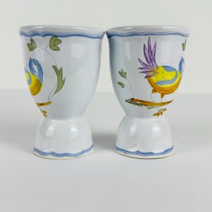 May include: Two white ceramic egg cups featuring a hand-painted design of colorful birds perched on branches. The birds have yellow bodies, blue wings, and purple tail feathers. Each cup has a scalloped base and a blue painted rim detail.