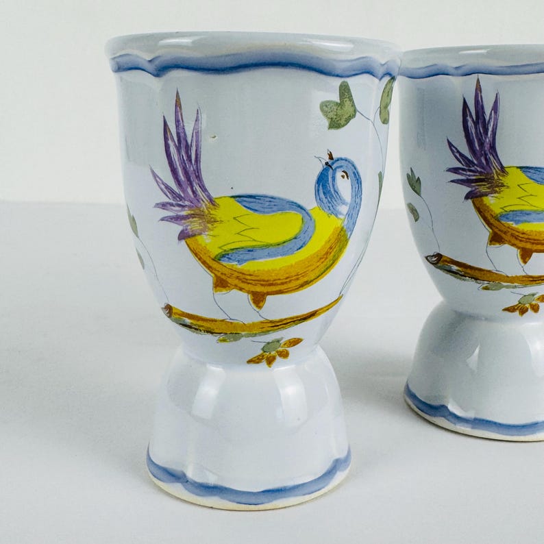 May include: Two white ceramic egg cups featuring a hand-painted design of a bird with yellow, blue, and purple plumage perched on a brown branch. The cups have a scalloped rim and a pedestal base, both accented with a blue painted wavy line border.