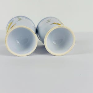 May include: Two light blue ceramic tumblers with a tapered base and a wide circular rim. The exterior surface features a subtle, abstract painted design in shades of yellow and grey, visible on the curved sides of the vessels.