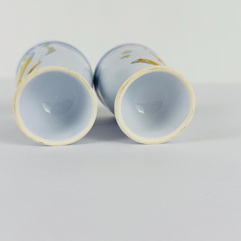 May include: Two light blue ceramic tumblers with a tapered base and a wide circular rim. The exterior surface features a subtle, abstract painted design in shades of yellow and grey, visible on the curved sides of the vessels.