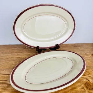 May include: Two off-white oval serving platters with a maroon rim and a maroon accent line. The platters are displayed on a wooden surface. The plates are a classic design for serving food.