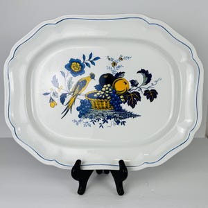 May include: White oval serving platter with a blue scalloped edge and a decorative fruit and bird design. The design features a yellow bird, blue grapes, and a basket of fruit. The platter is supported by a black stand.
