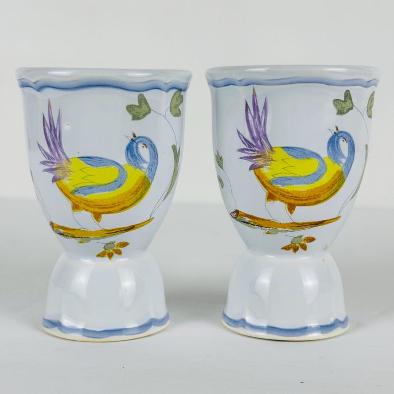 May include: Two white ceramic egg cups featuring a hand-painted design of a bird with a yellow body, blue wings, and purple tail feathers perched on a brown branch. The cups have a scalloped rim and base accented with a light blue painted trim.
