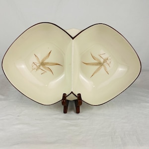 May include: A cream-colored ceramic divided serving dish with a dark brown rim. The dish features two symmetrical sections, each decorated with a brown botanical leaf illustration centered at the bottom of the bowl interior.