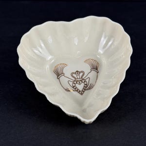 May include: A heart-shaped off-white ceramic dish with scalloped edges. The center features a brown line-art illustration of a Claddagh symbol, depicting two hands holding a crowned heart, rendered in a simple, clean, and minimalist graphic style.