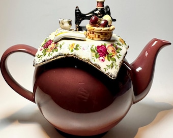 May include: A maroon ceramic teapot with a decorative lid featuring a miniature sewing machine, a basket of cherries, and a teacup. The lid has a floral pattern. The teapot has a curved handle and spout.