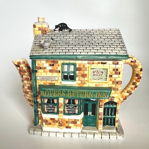 Op de afbeelding: Een keramische theepot in de vorm van een pub, met een bakstenen gevel, groene deuren en ramen en de tekst "Rovers Return Inn". Een zwart kattenbeeldje zit op het dak. De theepot heeft een handvat en een tuit en staat op een stenen voet.