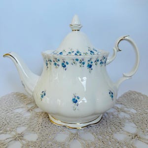 Puede incluir: Una tetera de porcelana blanca con tapa, adornada con pequeños motivos florales azules. La tetera tiene un pico curvo, un cuerpo redondeado y un ribete dorado. El asa también es blanca con detalles dorados. La tetera está sobre un tapete de encaje beige.