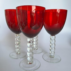 May include: Four red wine glasses. Each glass has a clear, bubble-style stem and a round base. The bowls are a deep, transparent red. The glasses are set on a white surface.