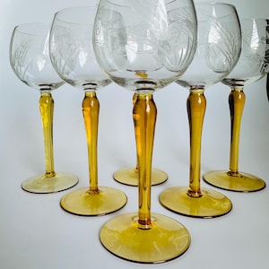 Op de afbeelding: Zes heldere wijnglazen met geëtste tarweontwerpen. De stelen en bases hebben een levendige amberkleur. De glazen zijn gerangschikt op een wit oppervlak, wat hun elegante ontwerp laat zien.