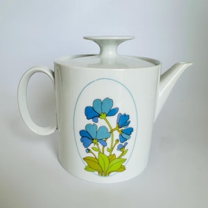 Peut inclure: Une théière en céramique blanche avec un corps cylindrique, une anse incurvée et un couvercle assorti doté d'un bouton sur pied. L'avant présente un contour ovale bleu contenant une illustration graphique de fleurs bleues, feuilles et tiges vertes.