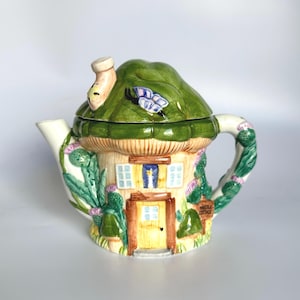 Może przedstawiać: Kapryśny ceramiczny imbryk w kształcie domku, z zielonym dachem, żółtymi drzwiami i kwiatowymi akcentami. Imbryk ma białą wylewkę i uchwyt, z napisem "Thistle Cottage" na znaku.