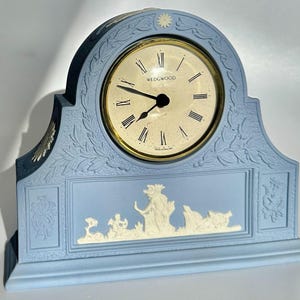 May include: A light blue Wedgwood mantel clock with a gold-tone clock face and Roman numerals. The clock features a decorative relief design with a central scene and floral accents. The clock measures approximately 10 inches wide.