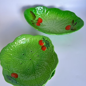 Puede incluir: Un juego de vajilla de cerámica con un diseño de hojas de col en verde vibrante, texturas en relieve y pequeños detalles de tomates rojos. La colección incluye un cuenco ovalado grande para servir y varios platos redondos con orificios.