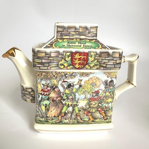 May include: A decorative ceramic teapot featuring a castle-shaped lid and a detailed illustration of Robin Hood in Sherwood Forest. The teapot has a cream-colored base with gold accents and a handle. The illustration includes figures in period clothing.