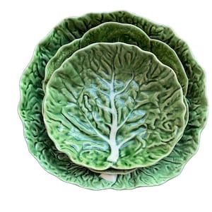 Puede incluir: Un juego de tres platos de cerámica apilables con un diseño de hoja de col verde texturizada. Cada plato tiene un borde festoneado y un patrón de nervadura central blanca en relieve que imita la apariencia de una hoja de col real.