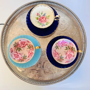 Op de afbeelding: Drie porseleinen theekopjes met schoteltjes op een rond, bewerkt zilverkleurig metalen dienblad. Twee sets hebben donkerblauwe schoteltjes met bloemmotief kopjes, terwijl de derde set een lichtblauw schoteltje en een roze bloemmotief kopje heeft.