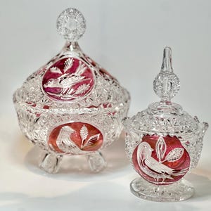 Puede incluir: Dos bomboneras de cristal transparente con tapas. Los platos tienen un interior rojo rubí y presentan diseños grabados de pájaros y hojas. El plato más grande tiene una base redonda con tres patas. El plato más pequeño tiene una base de pedestal.