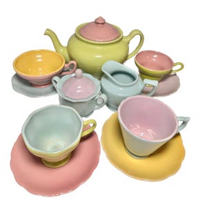 Op de afbeelding: Een pastelkleurige keramische theeset met een theepot, suikerpot, melkkan en verschillende kopjes en schotels. De set heeft verschillende kleuren, waaronder geel, roze en lichtgroen. De theepot heeft een roze deksel.