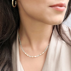 Puede incluir: Un delicado collar con pequeñas cuentas verde pálido intercaladas con perlas blancas y detalles dorados. Se ve un pendiente de aro dorado. La joya se lleva sobre un fondo de tono neutro.