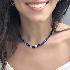 Puede incluir: Un collar de piedras preciosas azul oscuro con detalles plateados. El collar presenta piedras azul oscuro de forma irregular, pequeñas cuentas plateadas y dos cierres florales plateados. El collar se lleva alrededor del cuello.