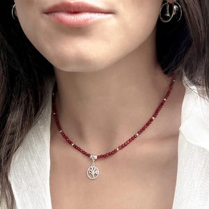 Puede incluir: Un collar de cuentas rojas con un colgante de árbol plateado. El collar está compuesto por pequeñas cuentas rojas redondas y cuentas plateadas. El colgante presenta un diseño detallado de árbol.