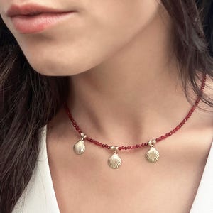 Puede incluir: Un delicado collar con una cadena de pequeñas cuentas rojas. Tres colgantes dorados en forma de concha marina están espaciados uniformemente a lo largo del collar. El collar se usa sobre una blusa blanca.