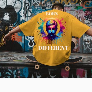 Op de afbeelding: Mosterdgeel T-shirt met een kleurrijke afbeelding op de rug. De afbeelding toont een gestileerd gezicht met rode ogen, omringd door een levendige explosie van kleuren. De woorden "BORN DIFFERENT" staan eronder gedrukt.